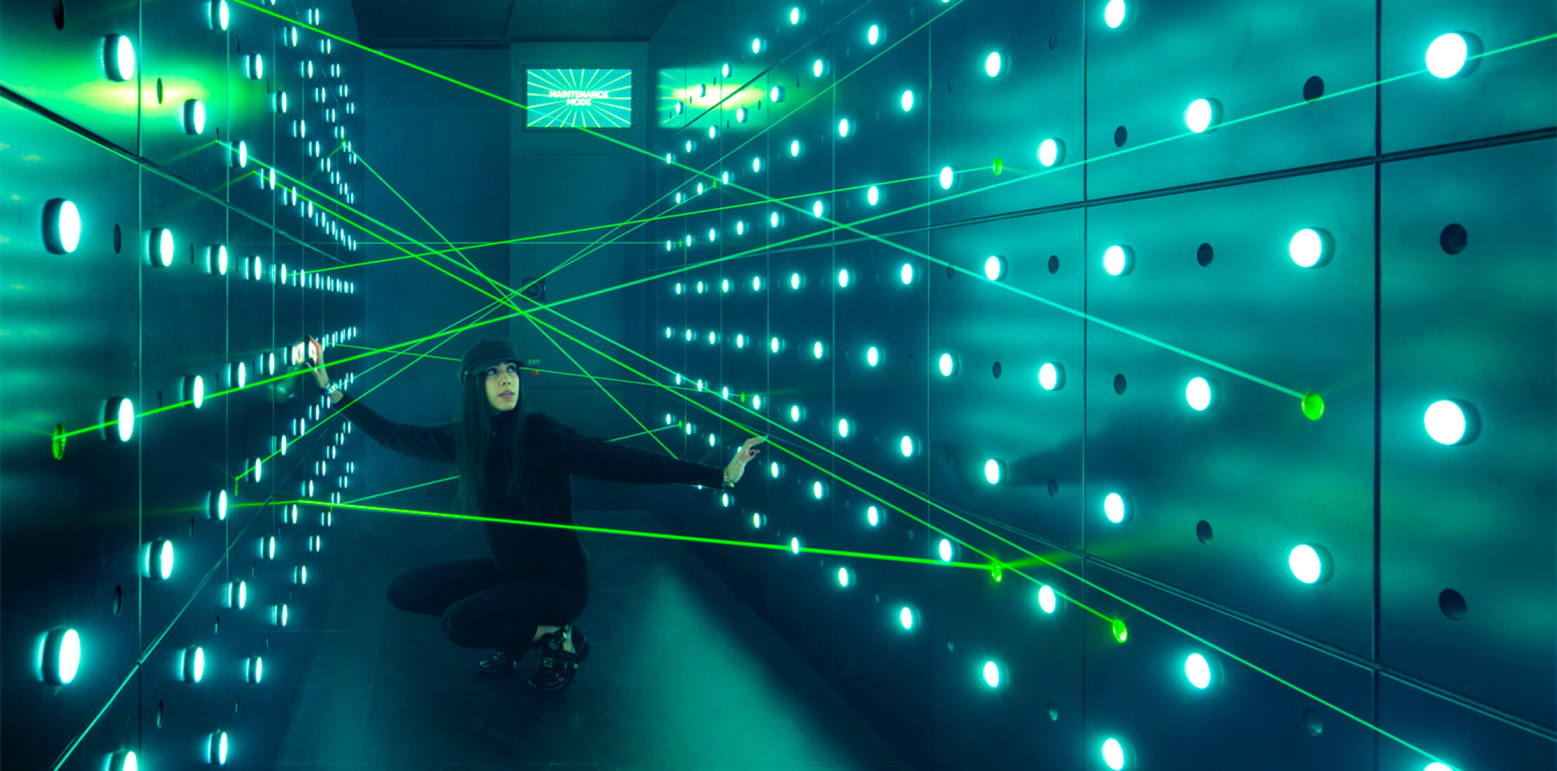 Colour image of a female visitor to the museum, crouching down inside a dark room surrounded by vivid green laser beams and light dots on the walls
