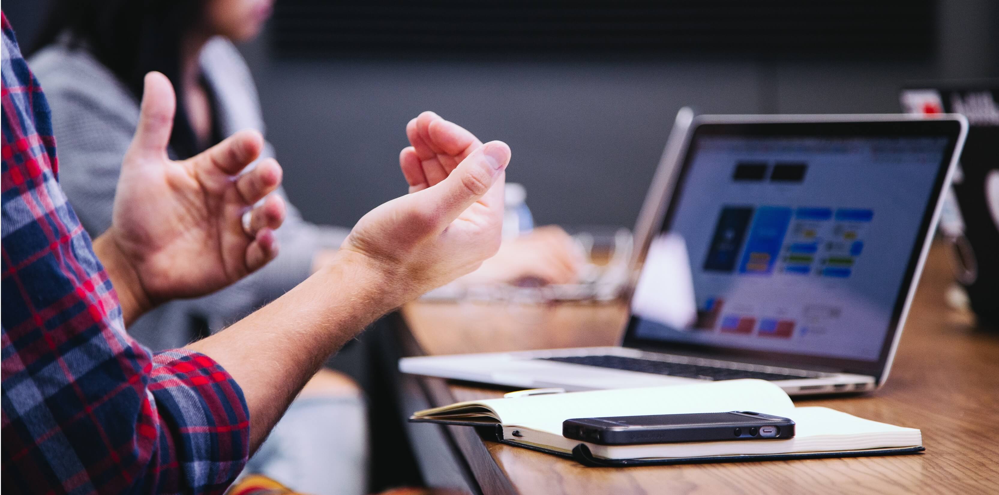 Close up shot of a man in a colourful checkered shirt, sat a table in a meeting room, discussing what is shown on his laptop screen